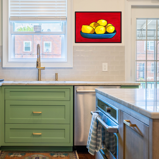 A black framed print of an oil painting of yellow lemons on a blue plate against a red background in a white kitchen with green cabinets