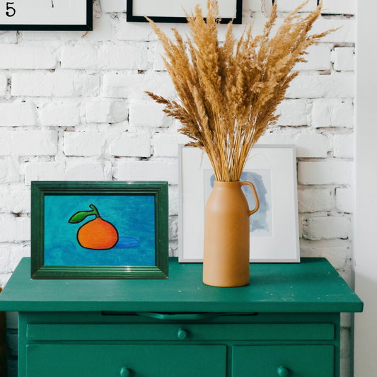 An oil painting of an orange clementine on a turquoise background in a vintage frame in a boho decor
