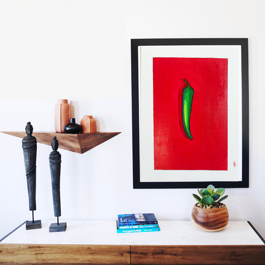 A framed oil painting of a green chili on a red background
