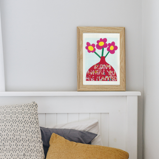 A wooden framed collage of three pink flowers with a yellow button in a red vase with the words bloom where you are planted