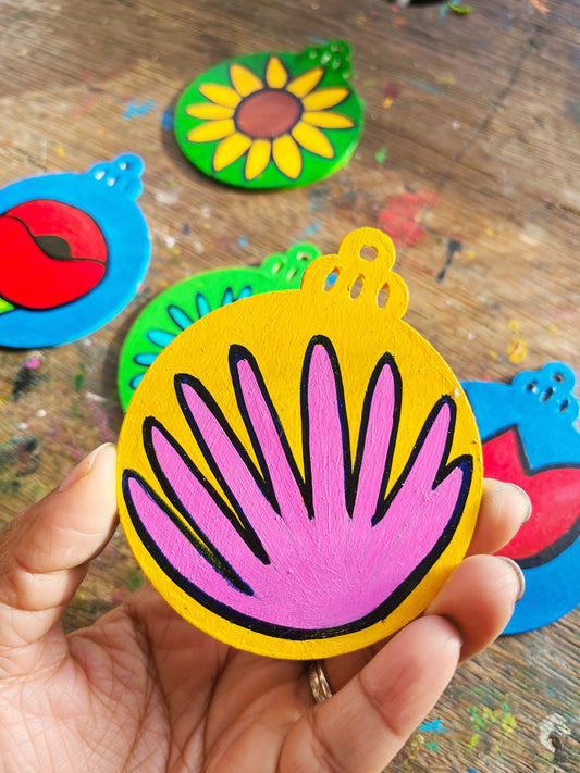 A hand holding a Christmas ornament representing a pink dahlia on a yellow background