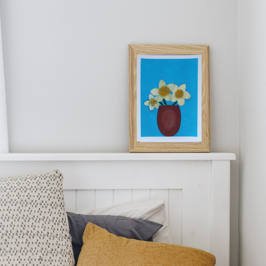 A wooden framed oil painting of white flowers in a brown pot against a blue background on a white chimney mantle