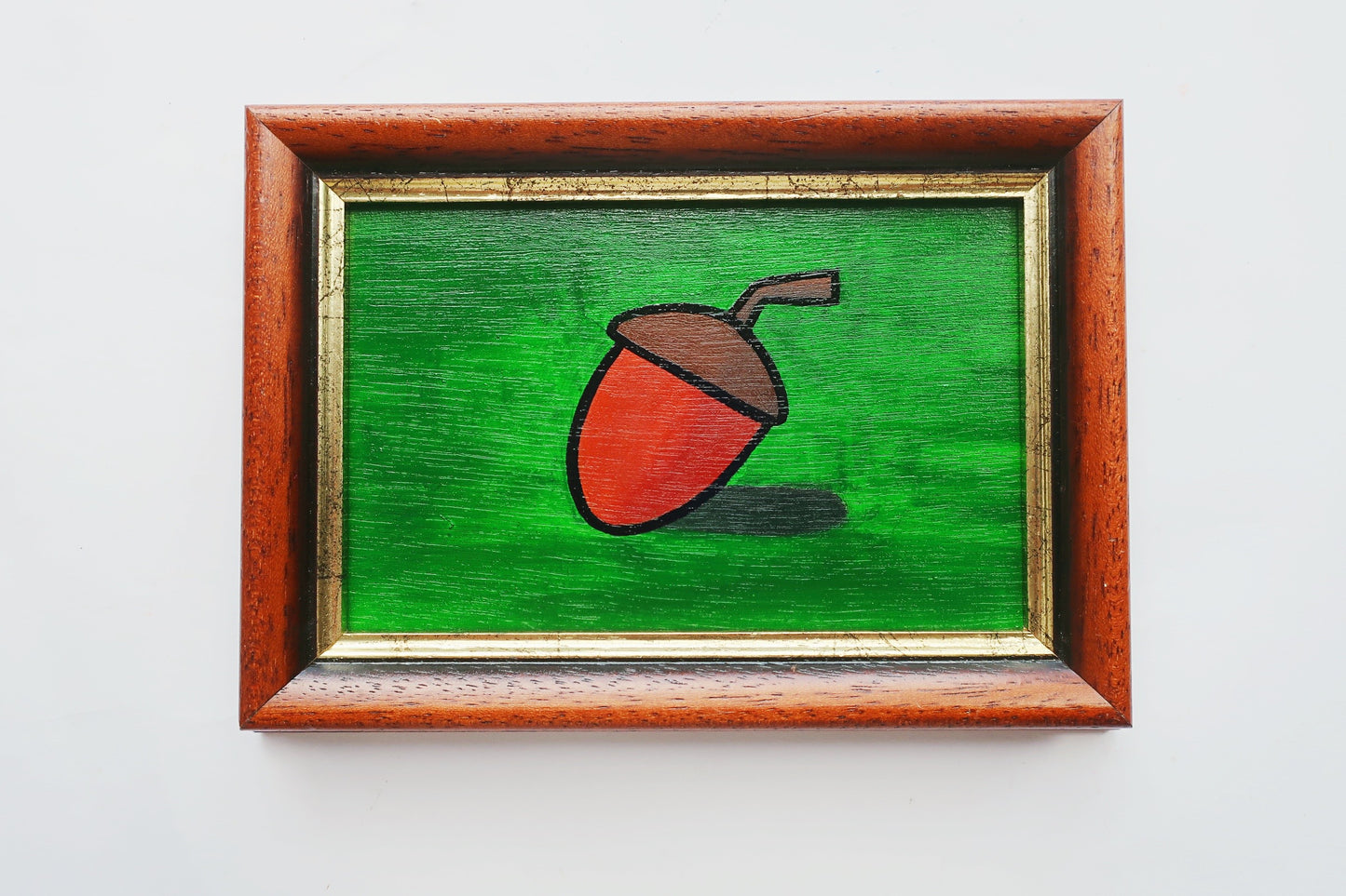 An oil painting of a brown acorn on a green background in a thrifted frame