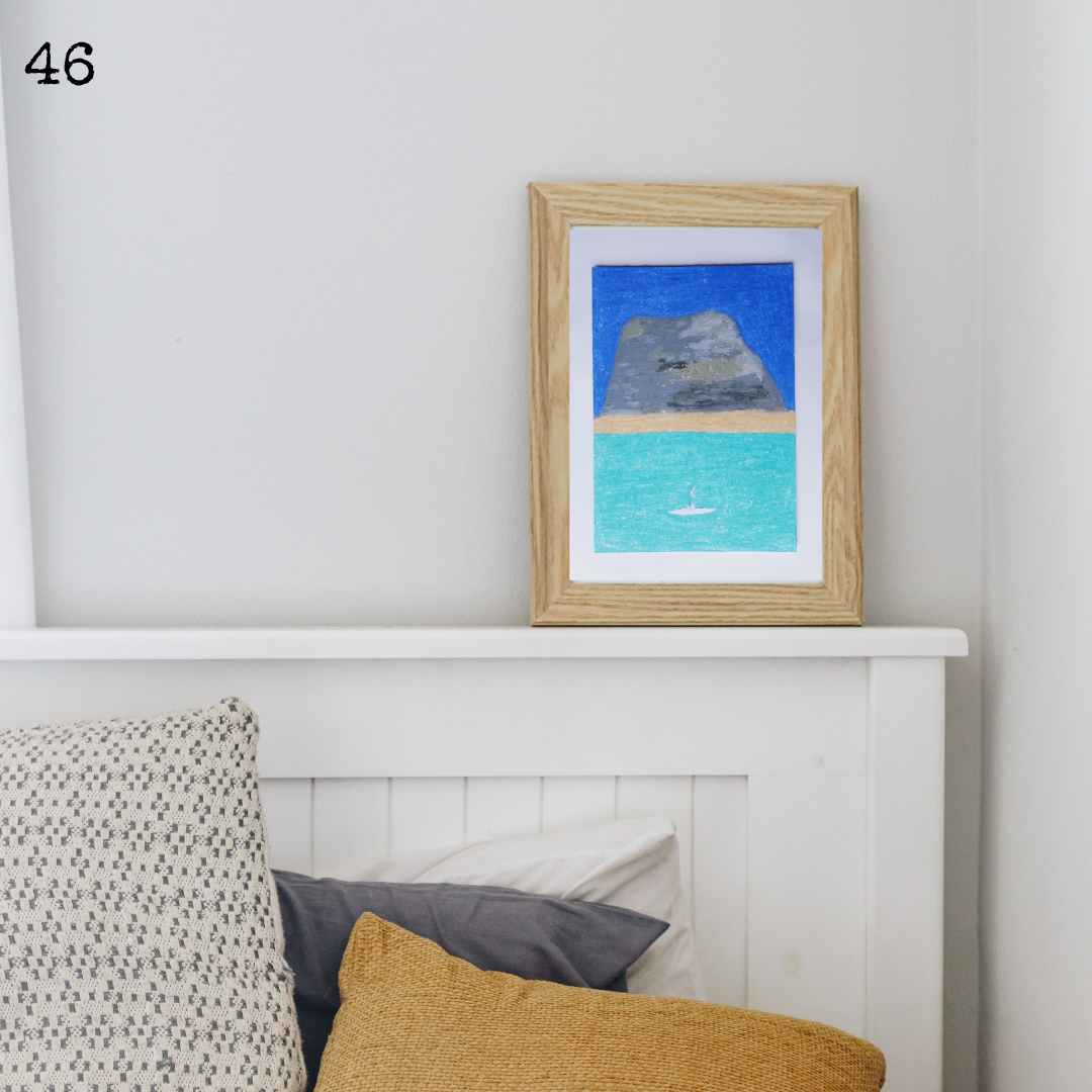 A wooden framed oil pastel painting of a beach in front of a mountain on a white mantelpiece