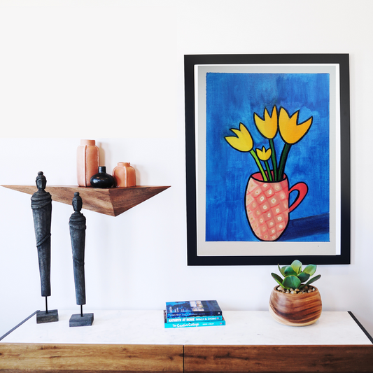 A black framed print of a printing of yellow tulips in a pink mug against a blue background